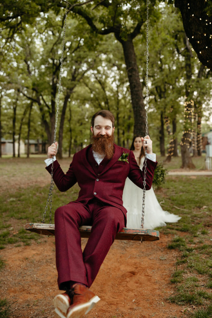 A groom in a bold maroon suit sits on a swing, with a bride in the background, creating a whimsical and romantic wedding scene
