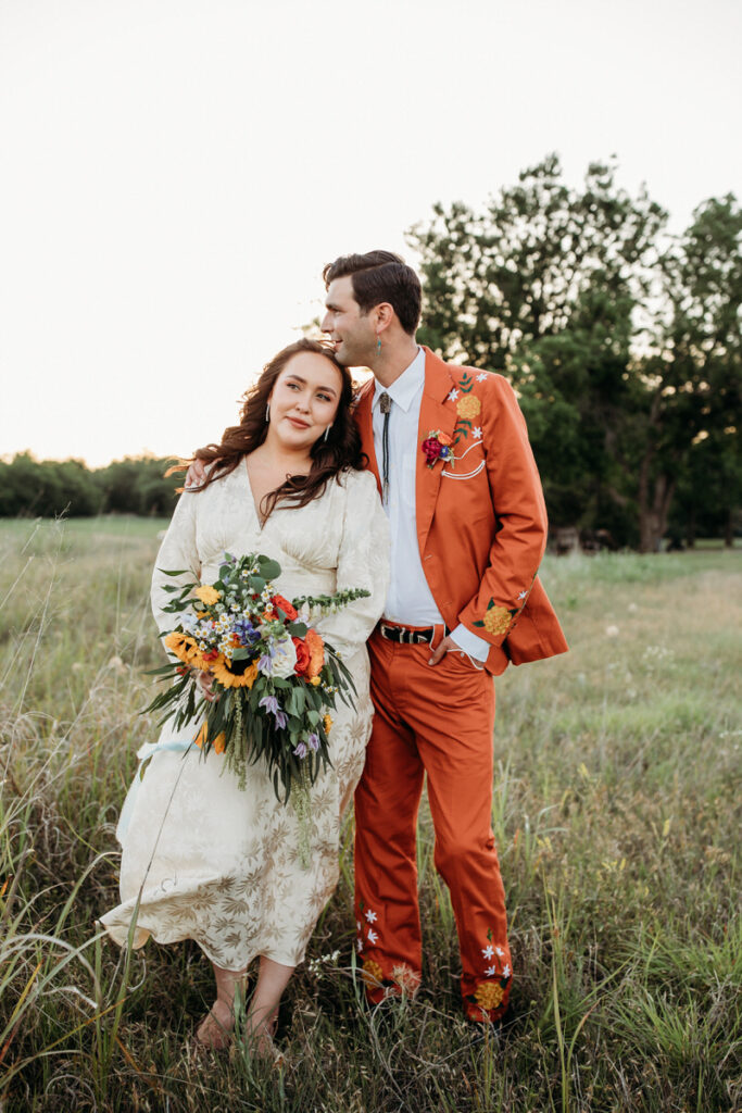 A couple stands together in a sunlit field, the man in an ornate orange suit and the woman in a vintage cream dress with a colorful bouquet