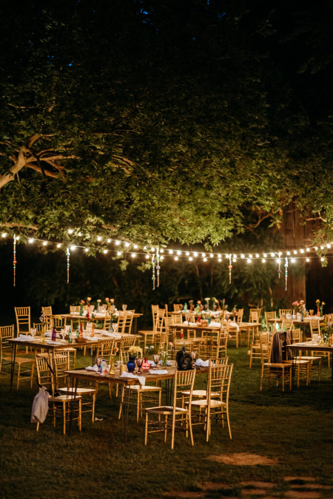 An enchanting evening reception set outdoors with tables arranged under a canopy of trees and string lights, offering a warm and intimate atmosphere.