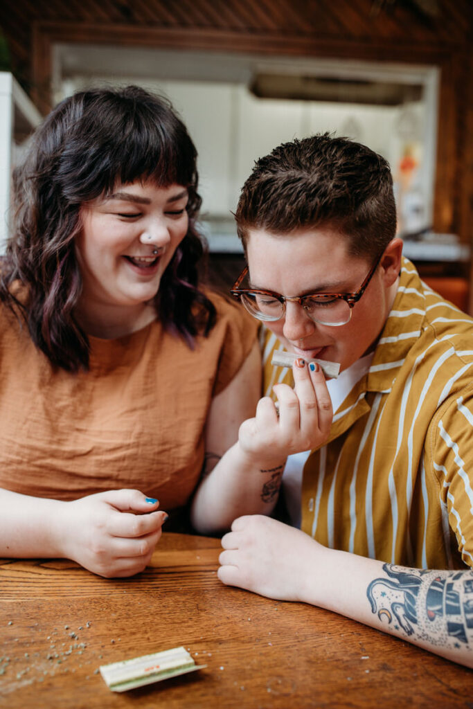 Lively at-home couple photoshoot featuring two individuals laughing together while preparing a joint, surrounded by cannabis paraphernalia on a wooden table
