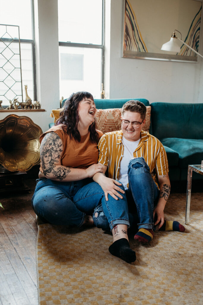 A couple in a home photoshoot laughing joyously, sitting on the floor with colorful socks, in a living room adorned with a teal couch and eclectic decor