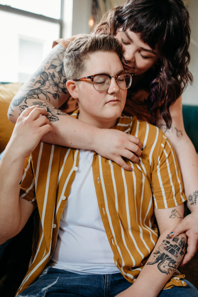 Intimate moment in a couple photoshoot at home, with one person embracing the other from behind, showcasing their tattoos in a warmly lit living room