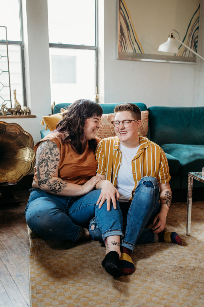 Casual and affectionate home photoshoot of a couple sitting on the floor, exchanging smiles against a backdrop of a teal couch and chic home decor