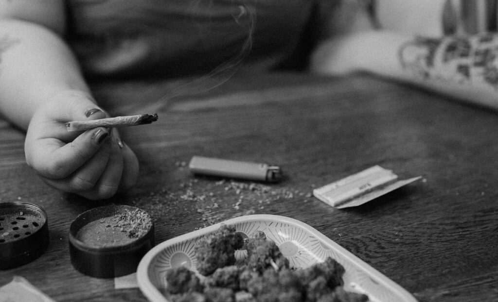 Artistic black and white photo of a person holding a lit joint with smoke wafting up, part of a relaxed couple photoshoot at home