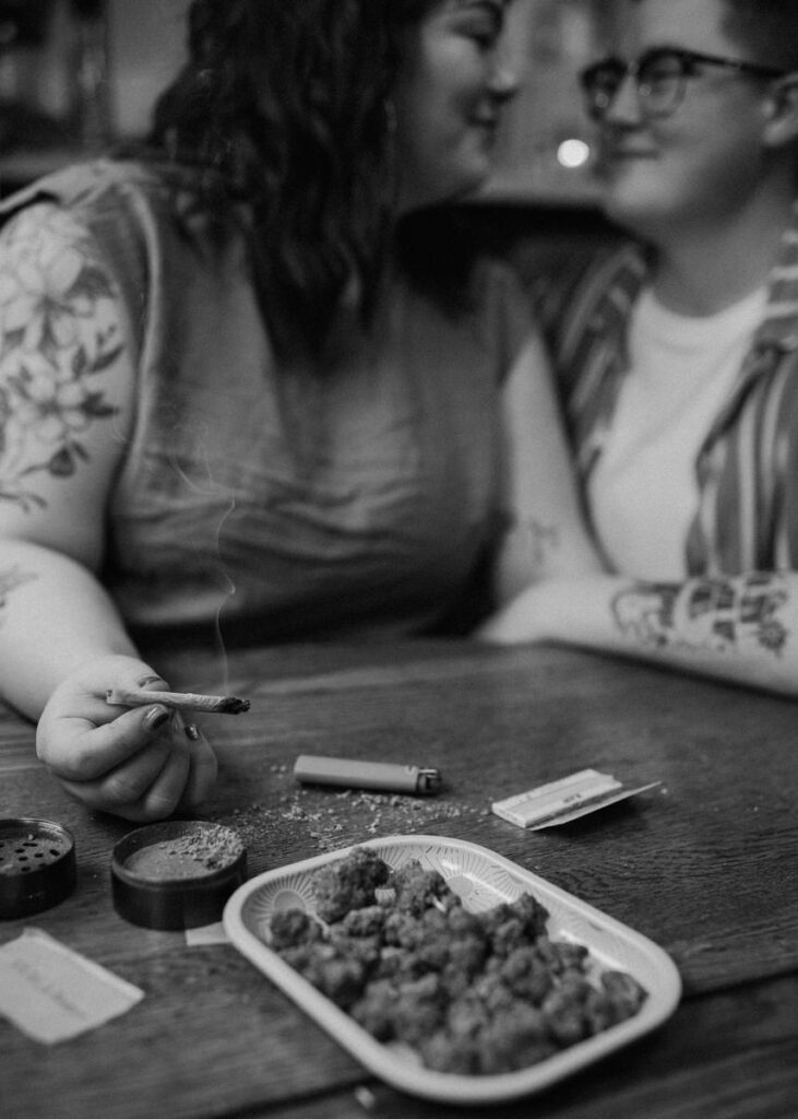 Couple engaging in a home photoshoot while preparing a joint, the table scattered with cannabis, a grinder, and a lighter, capturing a candid moment