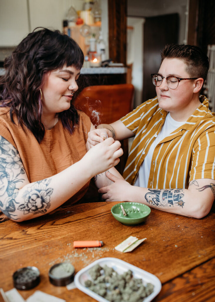 Couple in a home photoshoot affectionately lighting a joint together, with a focus on their hands and the smoke, in a cozy home setting