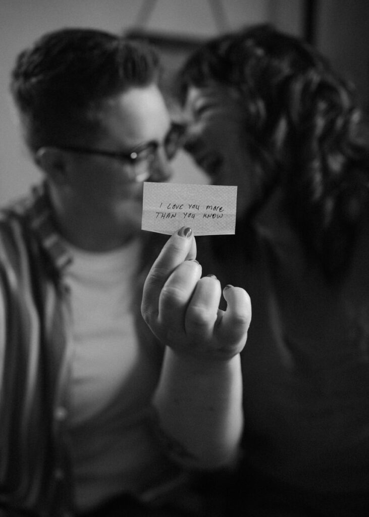 Black and white image capturing a poignant moment during a couple photoshoot at home, with a person holding a note saying 'I love you more than you know