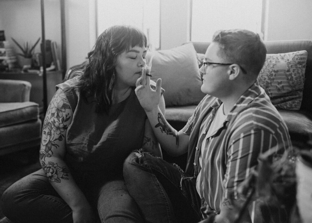 Black and white image of a couple's photoshoot at home, capturing an intimate moment as one partner gently places a joint to the other's lips in a serene setting