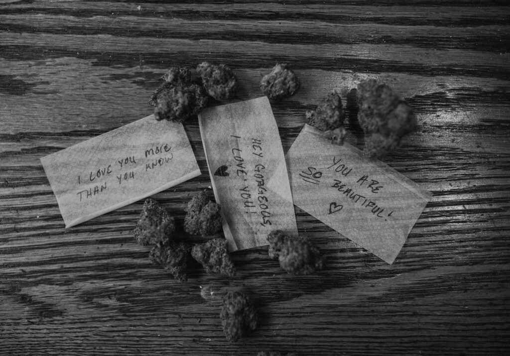 Close-up of cannabis buds and heartfelt, handwritten love notes scattered on a wooden surface, part of a couple's home photoshoot with a warm and affectionate vibe