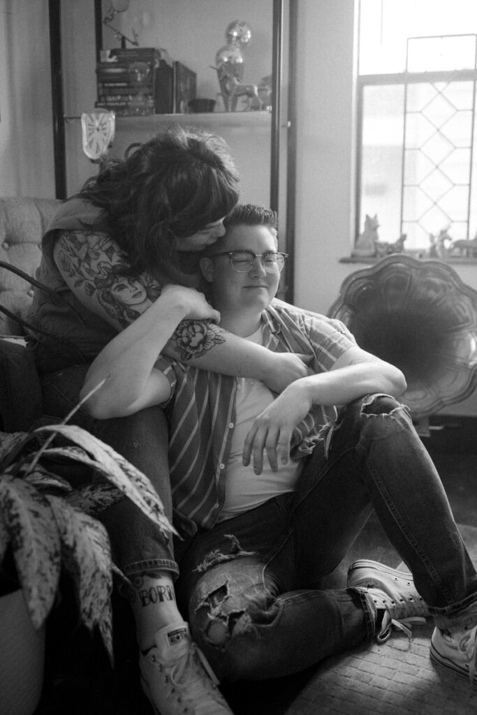 A tender monochrome snapshot of one partner embracing the other from behind, as they sit comfortably on a couch at home