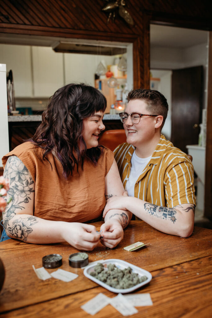 A couple engaged in a cozy home photoshoot, smiling as they sit closely together at a wooden table, with tattooed arms and a casual, affectionate atmosphere