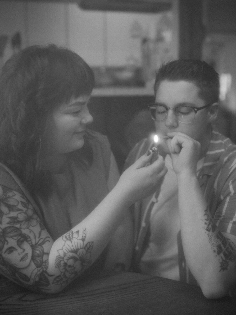 Black and white photo from a couple's intimate home photoshoot, highlighting a shared moment of lighting a joint, with a focus on their hands and the soft glow of the flame