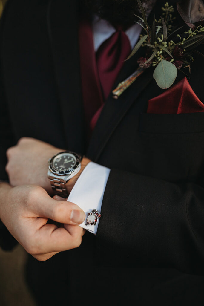 Close-up of a groom's hands adjusting his white shirt cuff, showcasing unique wedding cufflinks with a whimsical red and silver design