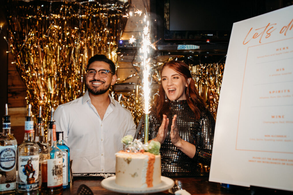 A beaming couple celebrating their wedding with a sparkler-lit cake in front of a shimmering gold tinsel backdrop, exuding a festive and glamorous vibe