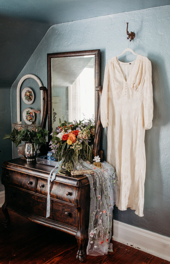 A vintage wedding dress hangs elegantly beside a floral arrangement and antique mirror, capturing a timeless bridal preparation scene