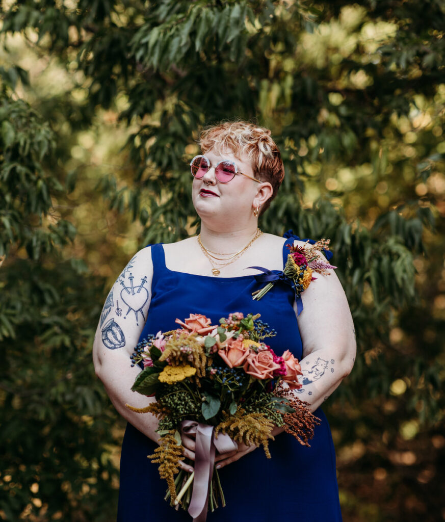 A wedding guest in a blue dress and sunglasses confidently poses with a vibrant bouquet, displaying her unique style and the colorful spirit of the event