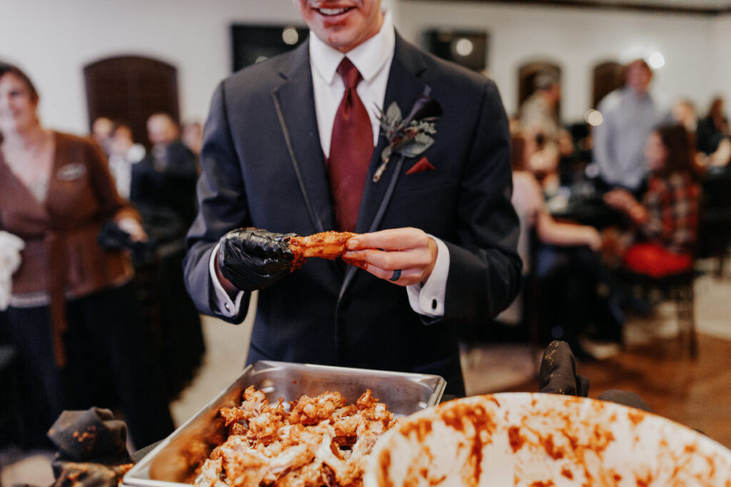 A groom in a dark suit participating in a hot wing eating contest during a wedding reception, with a tray of spicy wings in the foreground