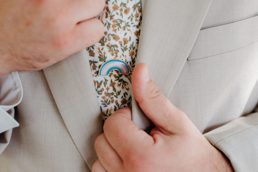 "A close-up of a groom's chest, with a lapel pin displaying the colors of the transgender flag on his floral tie, symbolizing inclusivity and pride