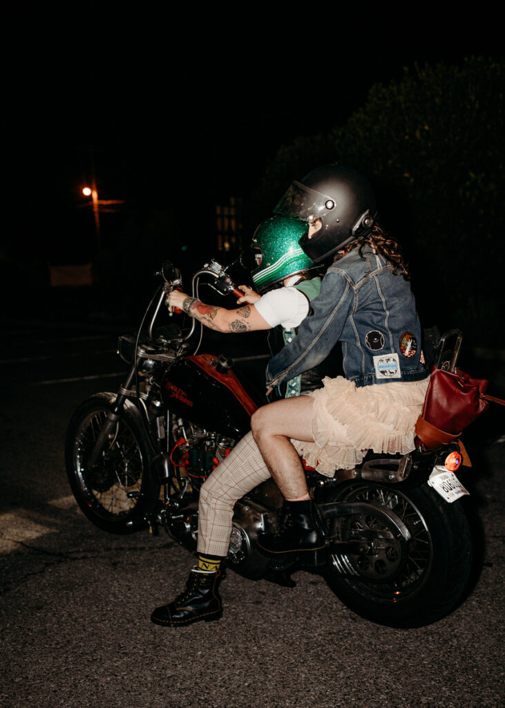 A couple in wedding attire riding a motorcycle at night, with the rider wearing a glittery green helmet and a denim jacket over a tulle skirt.