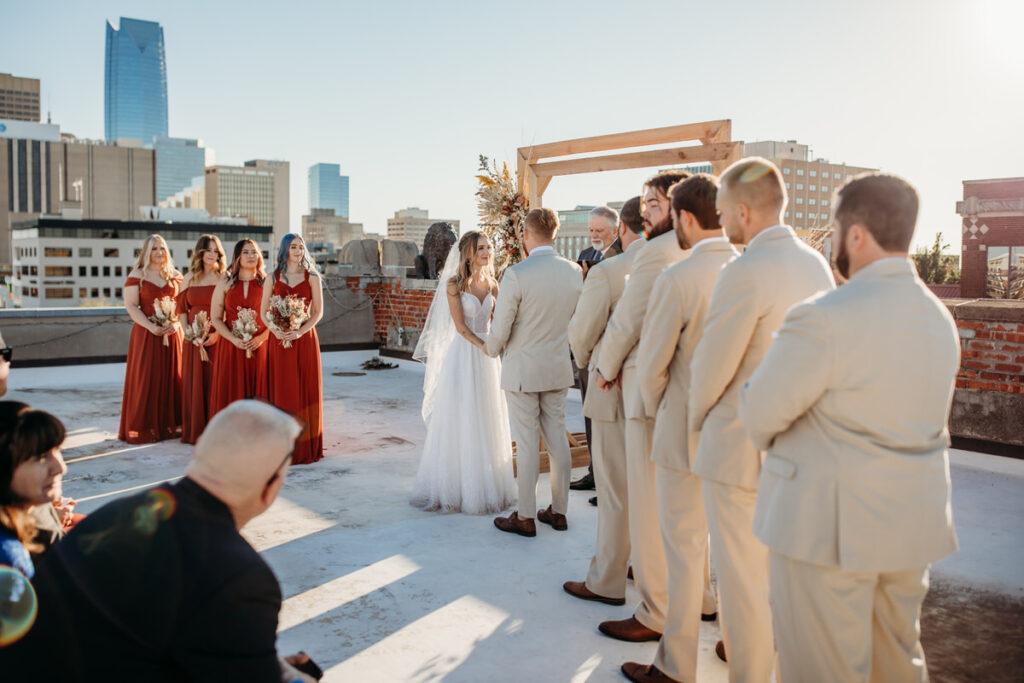 Bridal ceremony on a rooftop with a wooden arch, against the backdrop of Oklahoma City's skyline, showcasing the urban wedding venue