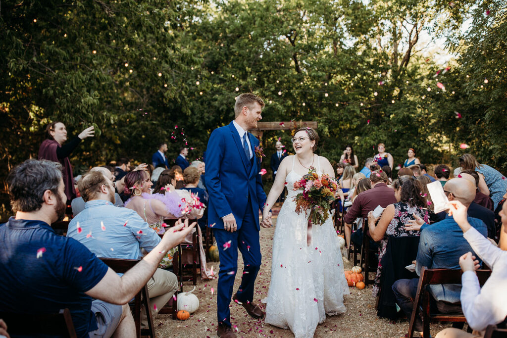 A bride and groom joyfully walking down the aisle as guests toss flower petals during their ceremony exit, set in an outdoor autumnal setting