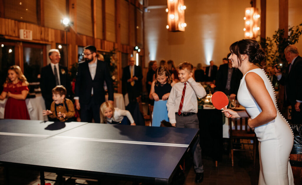 A lively wedding reception scene with guests, including children, playing a game of table tennis in a warmly lit hall.