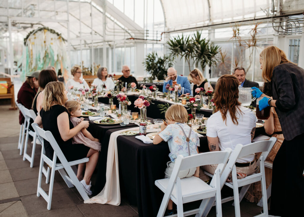 A relaxed and intimate garden dinner party at a wedding, with guests seated at a long table in a greenhouse setting, adorned with elegant floral centerpieces