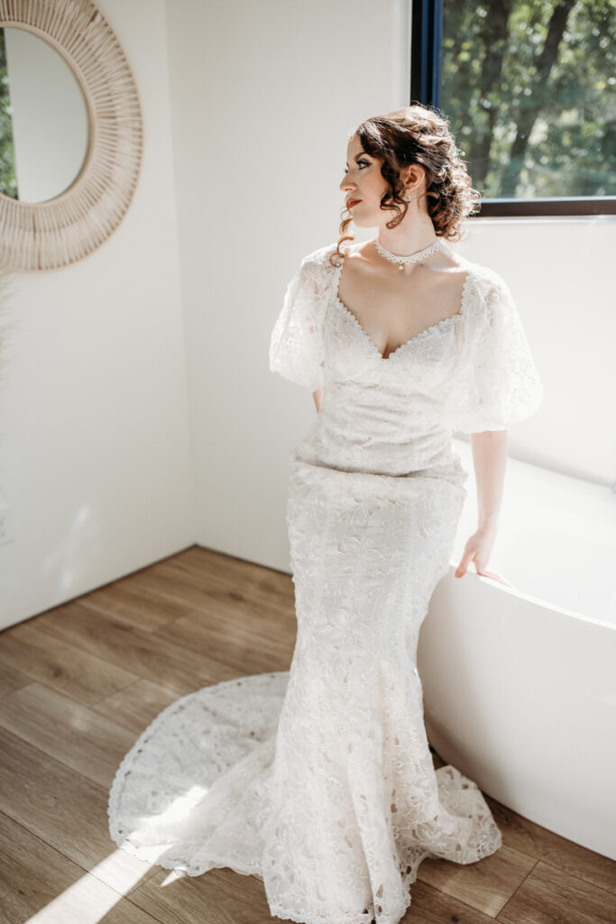 A bride gazes out a window, her silhouette accentuated by the intricate details of a vintage lace wedding gown with a dramatic neckline and delicate sleeves