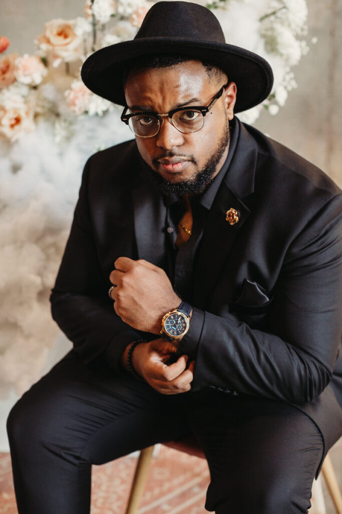 A stylish groom seated in a thoughtfully arranged setting, wearing a classic black suit and hat, his look completed with chic glasses and a luxury watch