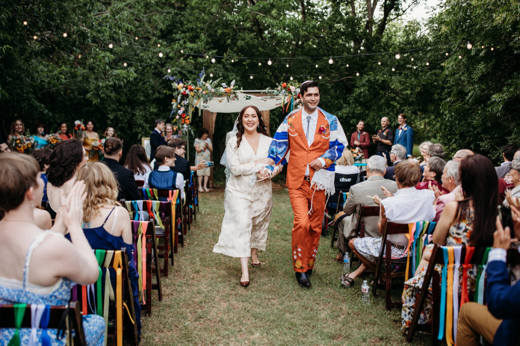 Joyful bride and groom walking down the aisle post-ceremony, with the bride in a white dress and the groom in a brightly colored, floral-patterned suit