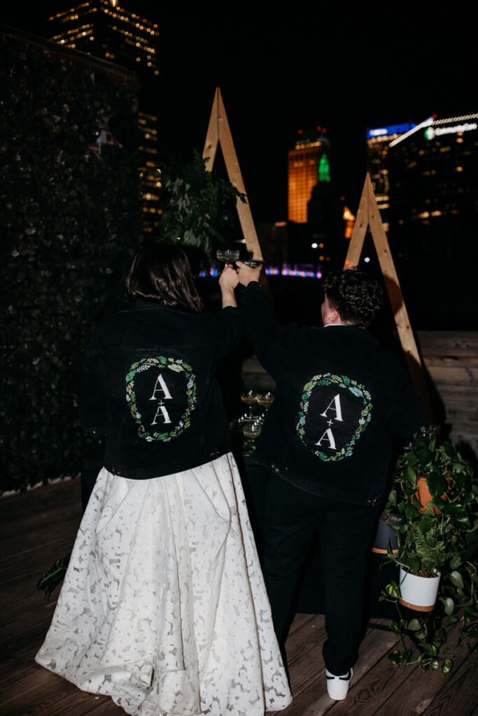 A couple from behind, wearing matching black jackets with personalized patches, holds a binoculars-like object while overlooking a cityscape at night