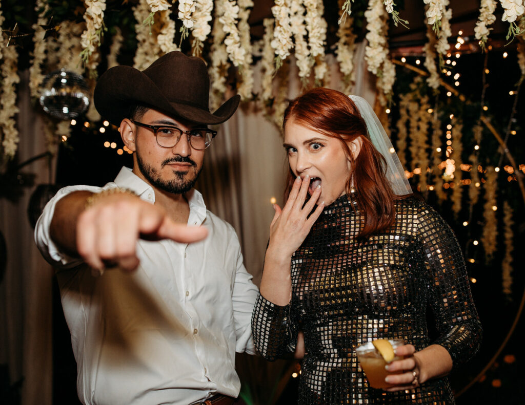 A bride and groom in a festive mood, the bride in a shimmering, patterned dress and the groom in a cowboy hat, pointing playfully at the camera