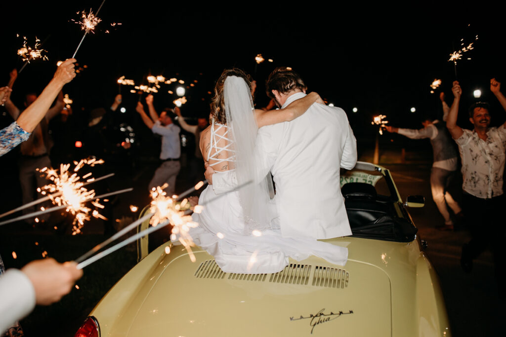 Newlyweds exiting their wedding in a vintage car, with guests holding sparklers on both sides, creating a sparkling archway