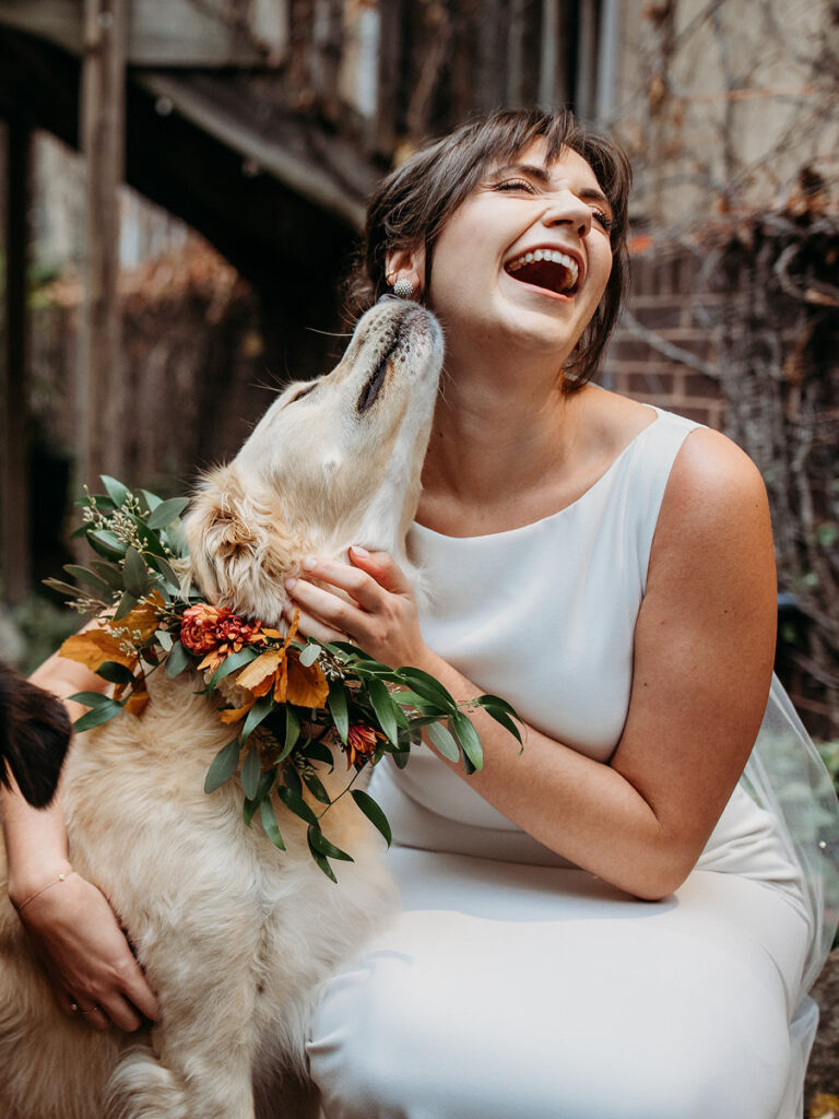 Bride laughing joyfully as she gets a loving kiss from her dog wearing a floral garland, highlighting a sweet and candid moment