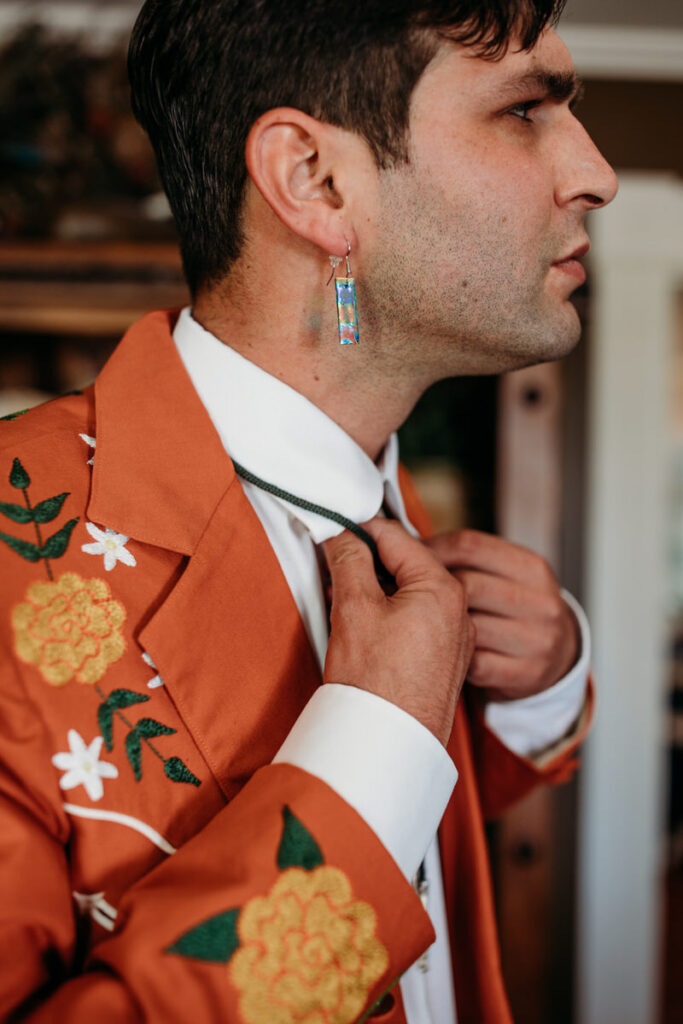 A groom adjusts his tie, wearing a western-inspired orange suit with intricate floral embroidery and a unique pair of long, colorful earrings