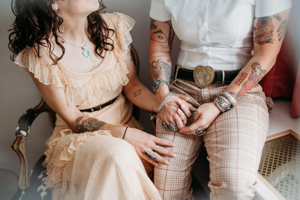 A couple seated close together, showcasing their unique styles with a sheer ruffled peach top and plaid trousers, surrounded by vintage decor